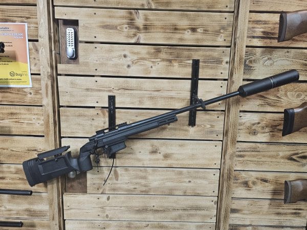Black sniper rifle with a suppressor mounted on a wooden display wall.