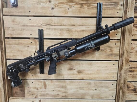 Black air gun with a carbon fiber barrel and a mounted air tank, displayed on a wooden wall.