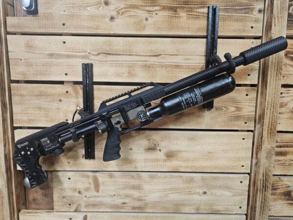 Black air gun with a carbon fiber barrel and a mounted air tank, displayed on a wooden wall.
